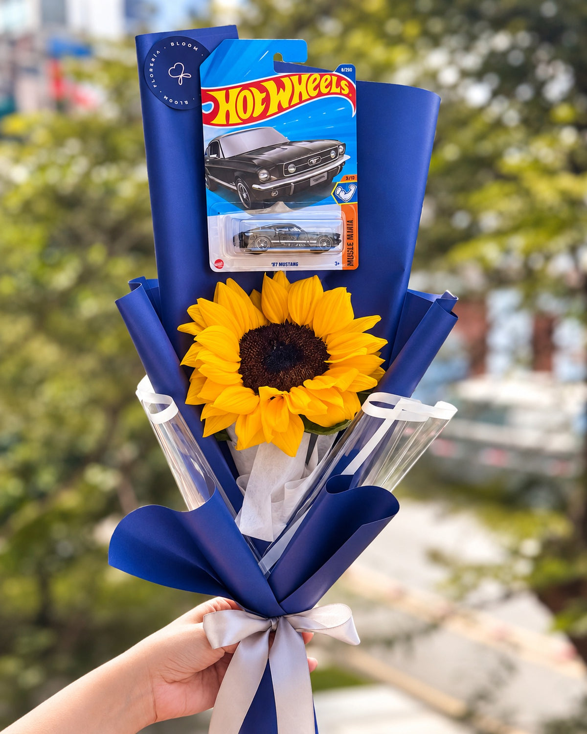 Ramo de girasol con Hot Wheels en envoltura azul y lazo plateado - Florería Bloom Lima